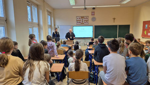 Zdjęcie przedstawia umundurowaną policjantkę i mężczyznę stojących przed siedzącymi uczniami.