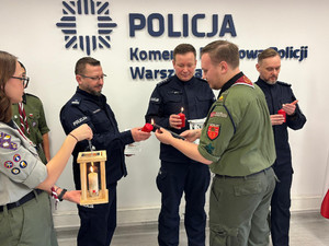 Na zdjęciu widać umundurowanych policjantów i harcerzy, którzy trzymają w dłoniach latarenki. Harcerz podpala świeczkę, którą trzyma w dłoniach policjant.