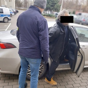 Zdjęcie przedstawia policjanta ubranego na ciemno, który ma odblaskową opaskę założoną na lewe przedramię i który umieszcza w samochodzie zatrzymanego mężczyznę ubranego na ciemno. Mężczyzna ten ma kajdanki założone na rękach trzymanych z tyłu. Część twarzy zasłania mu czarny prostokąt.