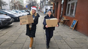 Zdjęcie przedstawia umundurowaną policjantkę i kobietę w czapce Mikołaja, które idą chodnikiem i niosą pudełka kartonowe.