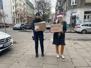 Zdjęcie przedstawia umundurowaną policjantkę i kobietę w czapce Mikołaja, które idą chodnikiem i niosą pudełka kartonowe.