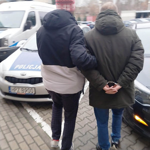 Na zdjęciu widoczny jest nieumundurowany policjant, który po swojej prawej stronie prowadzi osobę zatrzymaną ma kajdanki założone na rękach trzymanych z tyłu.