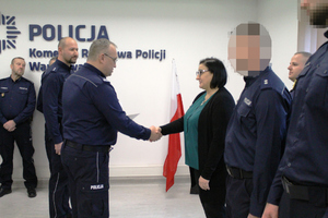 Zdjęcie przedstawia umundurowanych policjantów stojących na sali. Jeden z policjantów podaje rękę kobiecie ubranej w ciemny sweter i zieloną bluzkę.