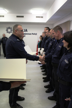 Zdjęcie przedstawia umundurowanych policjantów stojących na sali. Jeden z policjantów podaje rękę innemu policjantowi stojącemu w rzędzie wśród innych policjantów.