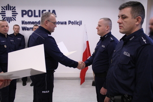 Zdjęcie przedstawia umundurowanych policjantów stojących na sali. Jeden z policjantów podaje rękę innemu policjantowi stojącemu w rzędzie wśród innych policjantów.