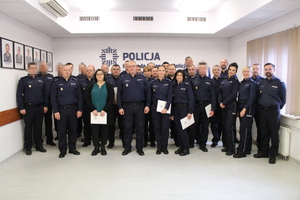 Zdjęcie przedstawia umundurowanych policjantów stojących na sali. Wszyscy patrzą w stronę obiektywu. Część z nich ma twarze zasłonięte komputerowym filtrem, niektórzy trzymają w dłoniach jasne papierowe teczki. Za ich plecami na ścianie znajduje się granatowy napis.