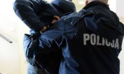 Zdjęcie przedstawi fragment sylwetki osoby ubranej w granatową kurtkę z białym napisem POLICJA, która przytrzymuje stojącą przed nim osobę.