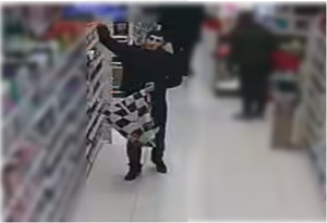 Zdjęcie przedstawia mężczyznę, który ma około 170 cm wzrostu, jest normalnej budowy ciała, ma krótkie czarne włosy, ubrany jest na ciemno. Trzyma w jednej ręce torbę biało-czarną, drugą sięga na półkę z asortymentem sklepowym.