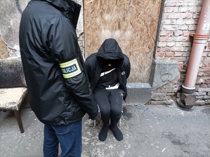 Zdjęcie przedstawia fragment sylwetki policjanta ubranego w ciemną kurtkę i opaskę z napisem policjanci na prawym przedramieniu, który stoi przy siedzącym na krześle ubranym na ciemno mężczyźnie w kapturze.