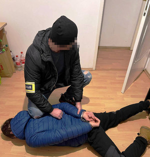 Zdjęcie przedstawia policjanta w cywilnych ubraniach, który ma na przedramieniu odblaskową opaskę z napisem POLICJA i pochyla się nad leżącym na ziemi mężczyzną w kajdankach.