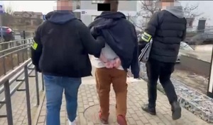 Zdjęcie przedstawia dwóch ubranych na ciemno mężczyzn z opaskami odblaskowymi na przedramionach, którzy prowadzą pomiędzy sobą zatrzymanego mężczyznę.