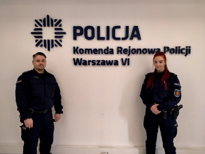 Zdjęcie przedstawia dwoje umundurowanych policjantów stojących na tle ściany z napisem Komenda Rejonowa Policji Warszawa VI.