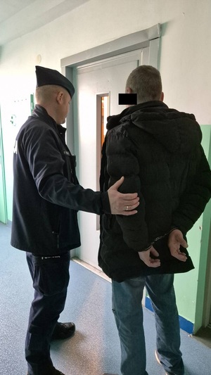 Zdjęcie przedstawia policjanta, który prowadzi po swojej prawej stronie mężczyznę ubranego w ciemną kurtkę. Mężczyzna ten ma kajdanki trzymane na rękach z tyłu.