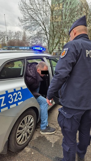 Zdjęcie przedstawia policjanta stojącego obok radiowozu do którego wsiada mężczyzna.