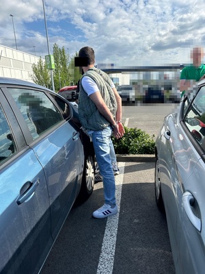 Zdjęcie przedstawia mężczyznę stojącego w kajdankach na rękach przy samochodzie, w tle widać stojącego policjanta.