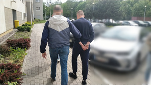 Zdjęcie przedstawia policjanta, który prowadzi po dziedzińcu, po swojej prawej stronie zatrzymanego mężczyznę. Mężczyzna ten ma kajdanki założone na rękach trzymanych z tyłu.