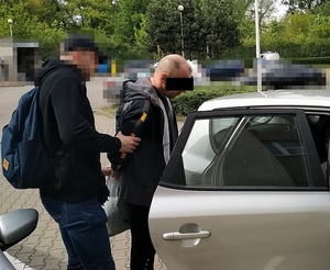Zdjęcie przestawia policjanta z zasłoniętą twarzą, który stoi za zatrzymanym mężczyzną i umieszcza go w samochodzie. Zatrzymany mężczyzna ma na części twarzy widoczny czarny prostokąt.