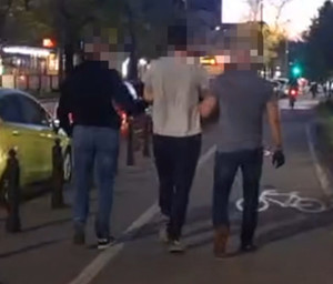 Zdjęcie przedstawia policjantów w ubraniach cywilnych, którzy prowadzą pomiędzy sobą chodnikiem zatrzymanego mężczyznę.