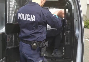 Zdjęcie przedstawia policjanta w mundurze, który ptrowadzi zatrzymanego mężczyznę.