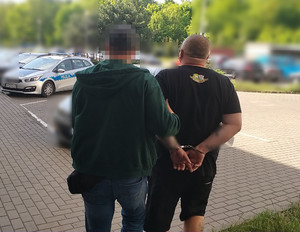Zdjęcie przedstawia ubranego na ciemno policjanta, który prowadzi po swojej prawej stronie zatrzymanego mężczyznę.