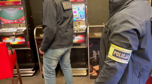 Zdjęcie przedstawia dwie sylwetki osób w kurtkach z napisem POLICJA, w tle widać automaty do gier.