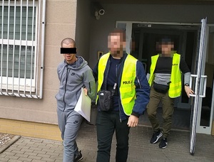 Zdjęcie przedstawia dwóch policjantów ubranych w odblaskowe kamizelki, którzy prowadzą przez dziedziniec zatrzymanego mężczyznę.