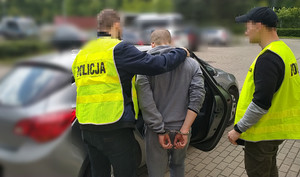 Zdjęcie przedstawia dwóch policjantów ubranych w odblaskowe kamizelki, którzy prowadzą przez dziedziniec zatrzymanego mężczyznę.