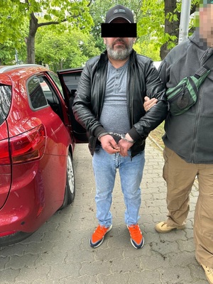Zdjęcie przedstawia sylwetkę mężczyzny stojącego przodem do zdjęcia z założonymi kajdankami. Obok niego stoi drugi mężczyzna, którego twarz jest zasłonięta komputerowym filtrem.