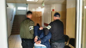 Zdjęcie przedstawia dwóch policjantów ubranych w cywilne ubrania i policyjne opaski odblaskowe na przedramionach, którzy prowadzą pomiędzy sobą przez korytarz zatrzymanego mężczyznę.