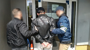 Zdjęcie przedstawia dwóch policjantów ubranych w cywilne ubrania i policyjne opaski odblaskowe na przedramionach, którzy wprowadzają do budynku zatrzymanego mężczyznę.