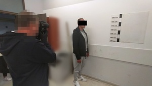 Zdjęcie przedstawia  sylwetki dwóch osób - policjant fotografuje mężczyznę stojącego przed jasną ścianą na tle policyjnej linijki do mierzenia wzrostu.