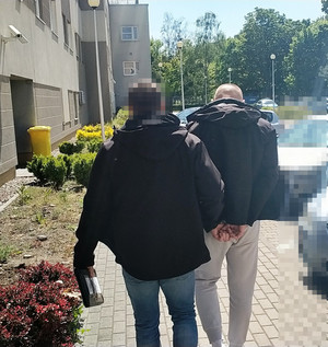 Zdjęcie przedstawia policjanta prowadzącego przez dziedziniec zatrzymanego mężczyznę.