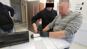 Zdjęcie przedstawia policjanta w czarnych ochronnych rękawiczkach, który przytrzymuje dłoń zatrzymanego mężczyzny nad kartką papieru.