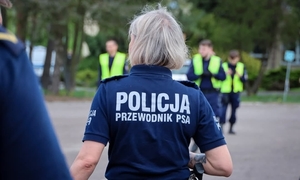 Zdjęcie przedstawia sylwetkę policjantki stojącej tyłem. Na jej koszulce widać napis POLICJA PRZEWODNIK PSA.