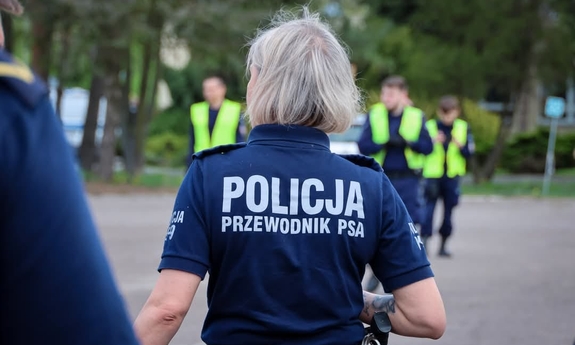Zdjęcie przedstawia sylwetkę policjantki stojącej tyłem. Na jej koszulce widać napis POLICJA PRZEWODNIK PSA.