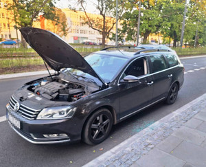 Zdjęcie przedstawia ciemnego volkswagena  z podniesioną maską i wybitą przednią szybą od strony kierowcy zaparkowanego przy ulicy.