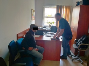 Zdjęcie przedstawia policjanta stojącego przy biurku, przy którym siedzi zatrzymany mężczyzna.