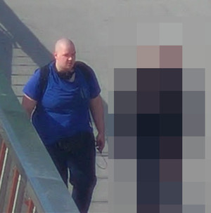 Zdjęcie przedstawia mężczyznę idącego przy barierce. Jest on w wieku 25-35 lat. Mężczyzna jest łysy, bez widocznego zarostu na twarzy. Ubrany jest w niebieską koszulkę z krótkim rękawem, i ciemne spodnie.