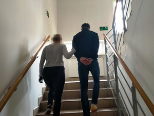 Zdjęcie przedstawia policjantkę w ubraniach cywilnych, która wprowadza po schodach mężczyznę w kajdankach na rękach trzymanych za plecami.