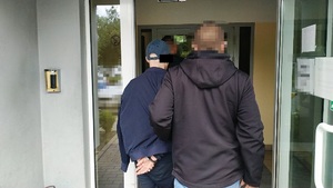 Zdjęcie przedstawia dwóch policjantów w ubraniach cywilnych, którzy wprowadzają do budynku zatrzymanego mężczyznę ubranego w czapkę z daszkiem.
