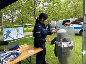 Zdjęcie przedstawia policjantkę stojącą obok dziecka ukrytego za policyjną tarczą.