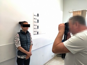 Zdjęcie przedstawia policjanta wykonującego zdjęcie kobiecie stojącej pod jasną ścianą z miarką do wzrostu.