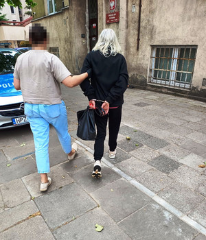 Zdjęcie przedstawia policjantkę prowadzącą po chodniku kobietę w kajdankach.