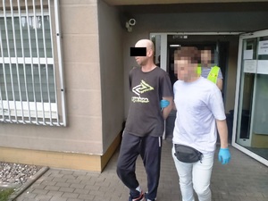 Zdjęcie przedstawia policjanta wyprowadzającego z budynku komendy zatrzymanego mężczyznę.