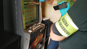 Zdjęcie przedstawia fragment sylwetki osoby z odblaskową opaską na przedramieniu stojącej obok automat do gier hazardowych.