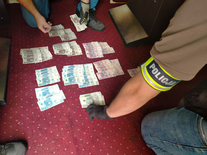 Zdjęcie przedstawia fragment sylwetki osoby, z odblaskową opaską na przedramieniu &quot;POLICJA&quot; pochylającej się nad rozłożonymi na wykładzinie banknotami.