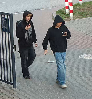 Zdjęcie przedstawia idące chodnikiem dwie młode osoby ubrane w ciemne bluzy z kapturami założonymi na głowach.