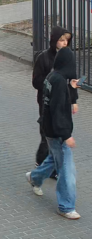Zdjęcie przedstawia idące chodnikiem dwie osoby ubrane w ciemne bluzy z kapturami założonymi na głowach.