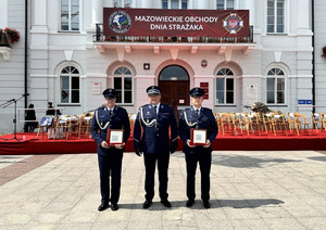 Zdjęcie przedstawia trzech umundurowanych galowo policjantów stojących w jednym rzędzie na tle napisu Mazowieckie Obchody Dnia Strażaka.