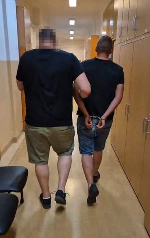 Zdjęcie przedstawia policjanta prowadzącego przez korytarz, po swojej prawej stronie zatrzymanego mężczyznę.